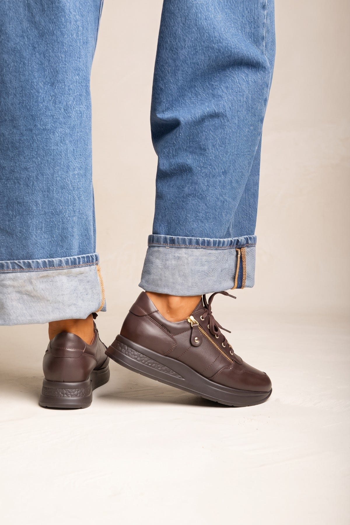 Model legs in cuffed jeans with brown leather lace up wedge sneakers side by side showing the raised thick sole