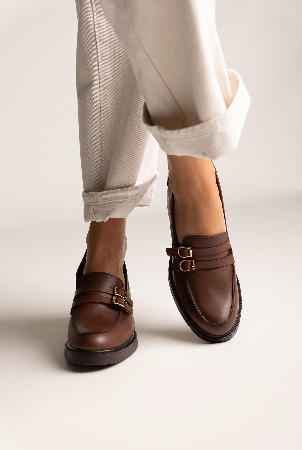 Brown leather loafers with double buckle straps in gold, rounded toe and low heel, worn on model, front view