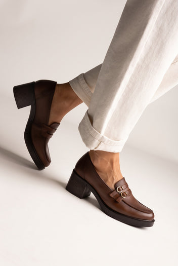 Brown leather block heel loafers with double gold buckle straps and platform sole, legs crossed, worn on model