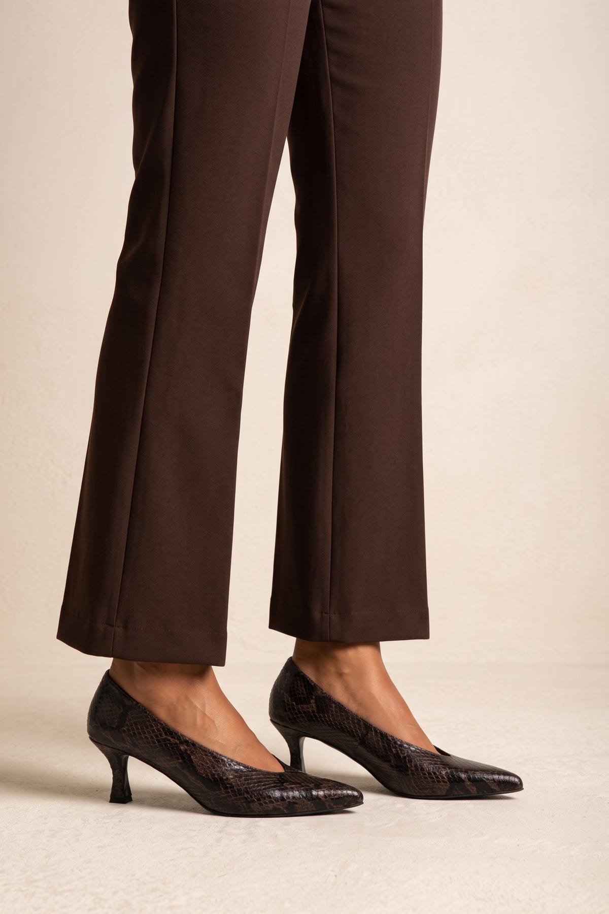Model with brown trousers wearing brown snake-print pointed pumps, side view of the feet and heel