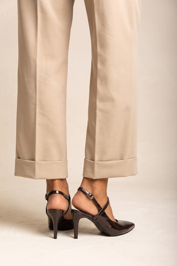 Lower legs of a model in beige cuffed trousers wearing brown patent slingback pumps with crossed straps