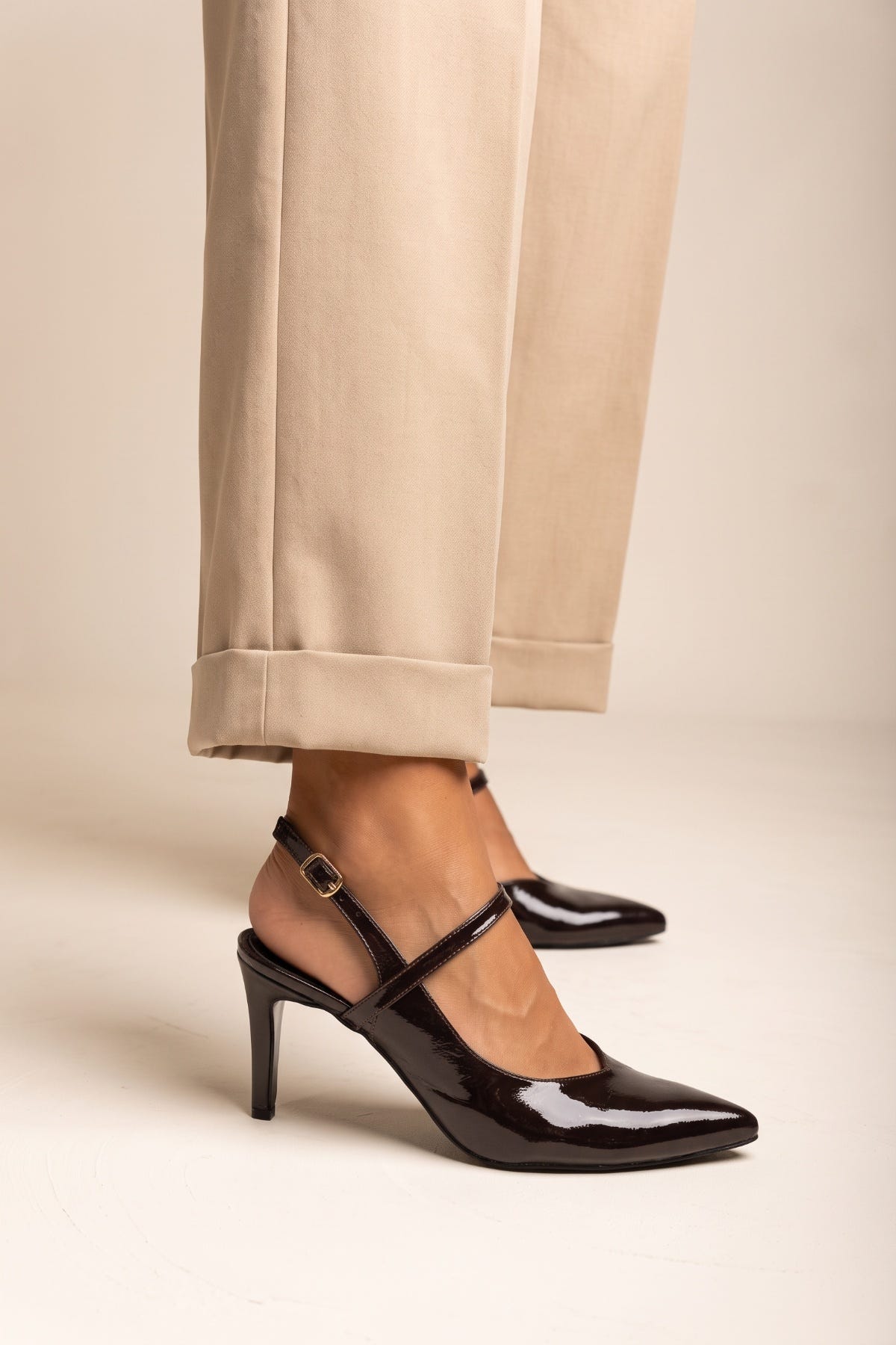 Model in beige trousers wearing brown patent slingback pumps with crossed straps seen in side view