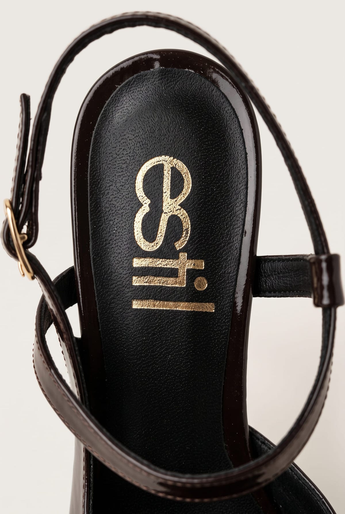 Close-up of a brown patent ankle strap and buckle beside a gold emblem on a black insole
