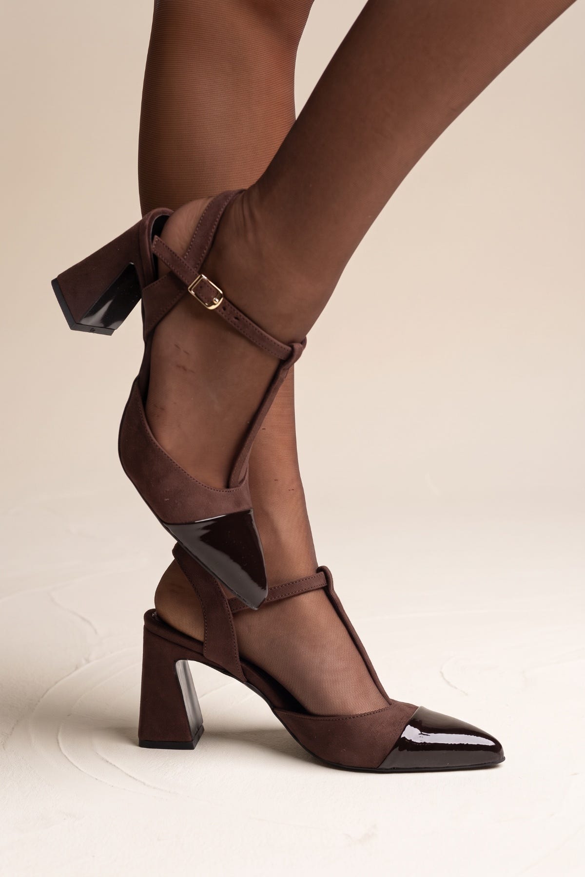 Model with leg raised showing chocolate suede T-strap block heel pump with black patent toe cap and buckle