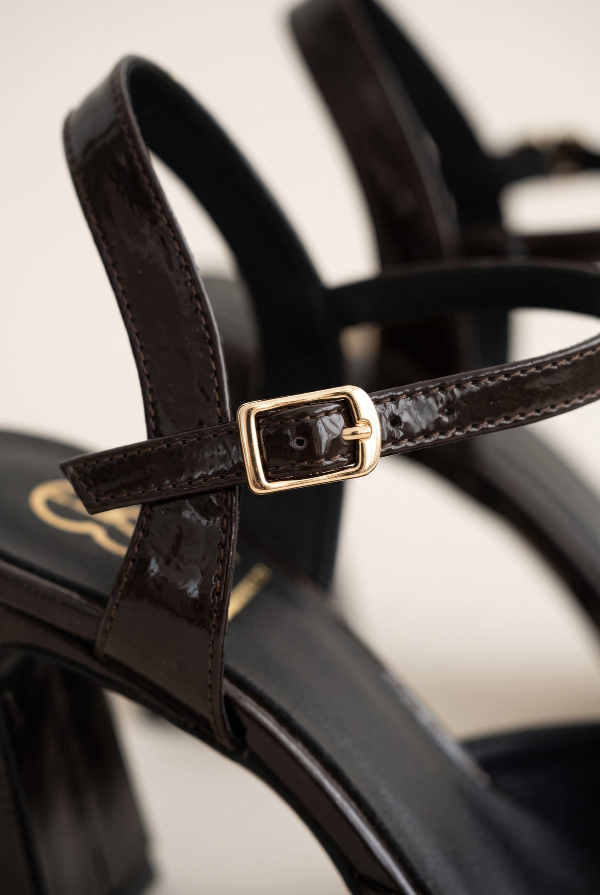 Close-up of brown patent T-strap showing ankle strap, gold-tone buckle and glossy leather finish