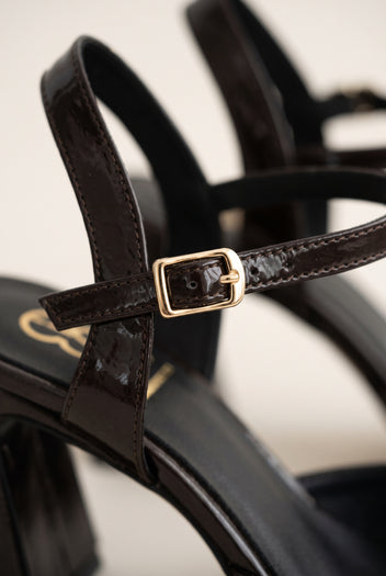 Close-up of brown patent T-strap showing ankle strap, gold-tone buckle and glossy leather finish