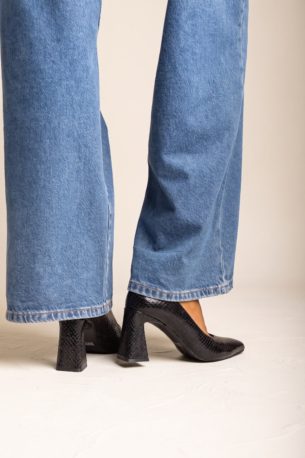Black snake-embossed pumps with block heel and pointed toe, front view of both feet in wide-leg jeans
