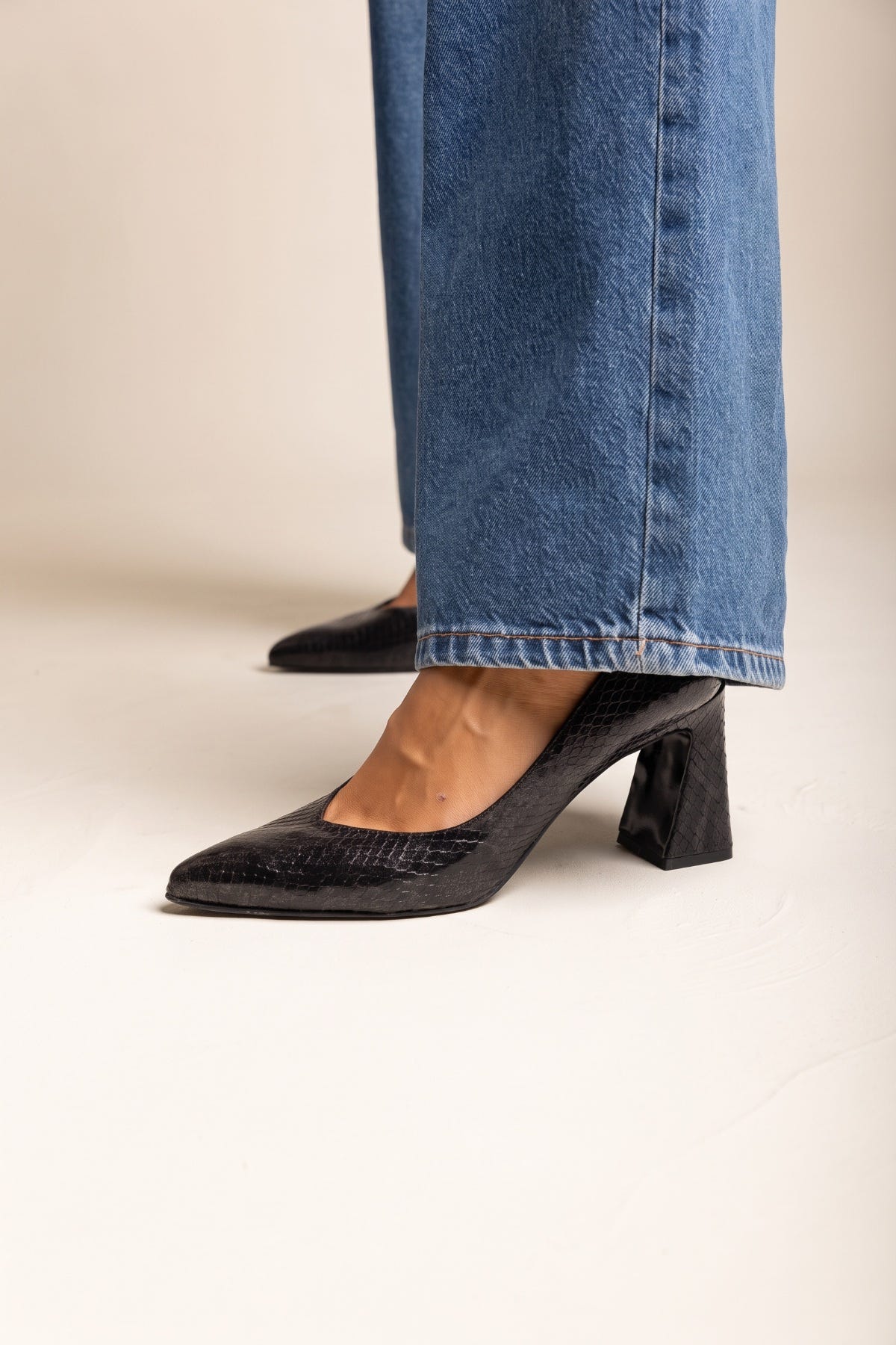 Black snake-embossed pointed toe pumps with block heel, side view of both feet in blue jeans