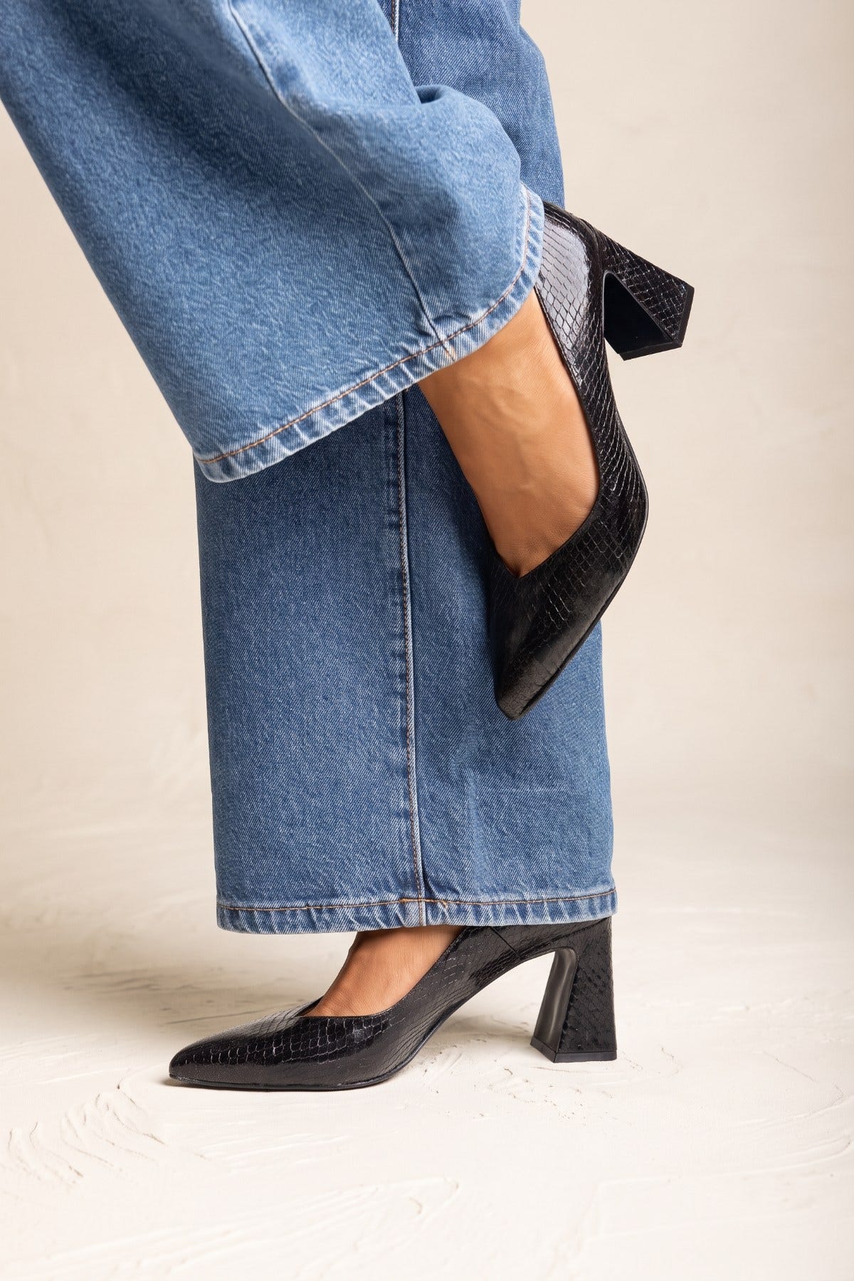 Model lifting leg to show black snake-embossed pointed toe pump with block heel, close side view with denim jeans