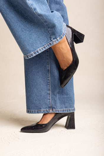 Model lifting leg to show black snake-embossed pointed toe pump with block heel, close side view with denim jeans