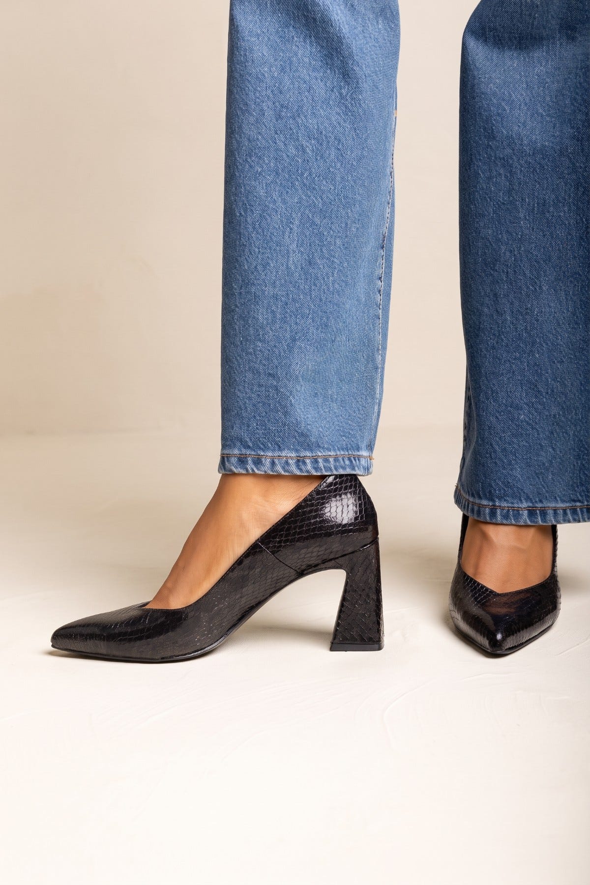 Model wearing black snake-embossed pointed toe block heel pumps, side view with light blue wide-leg jeans
