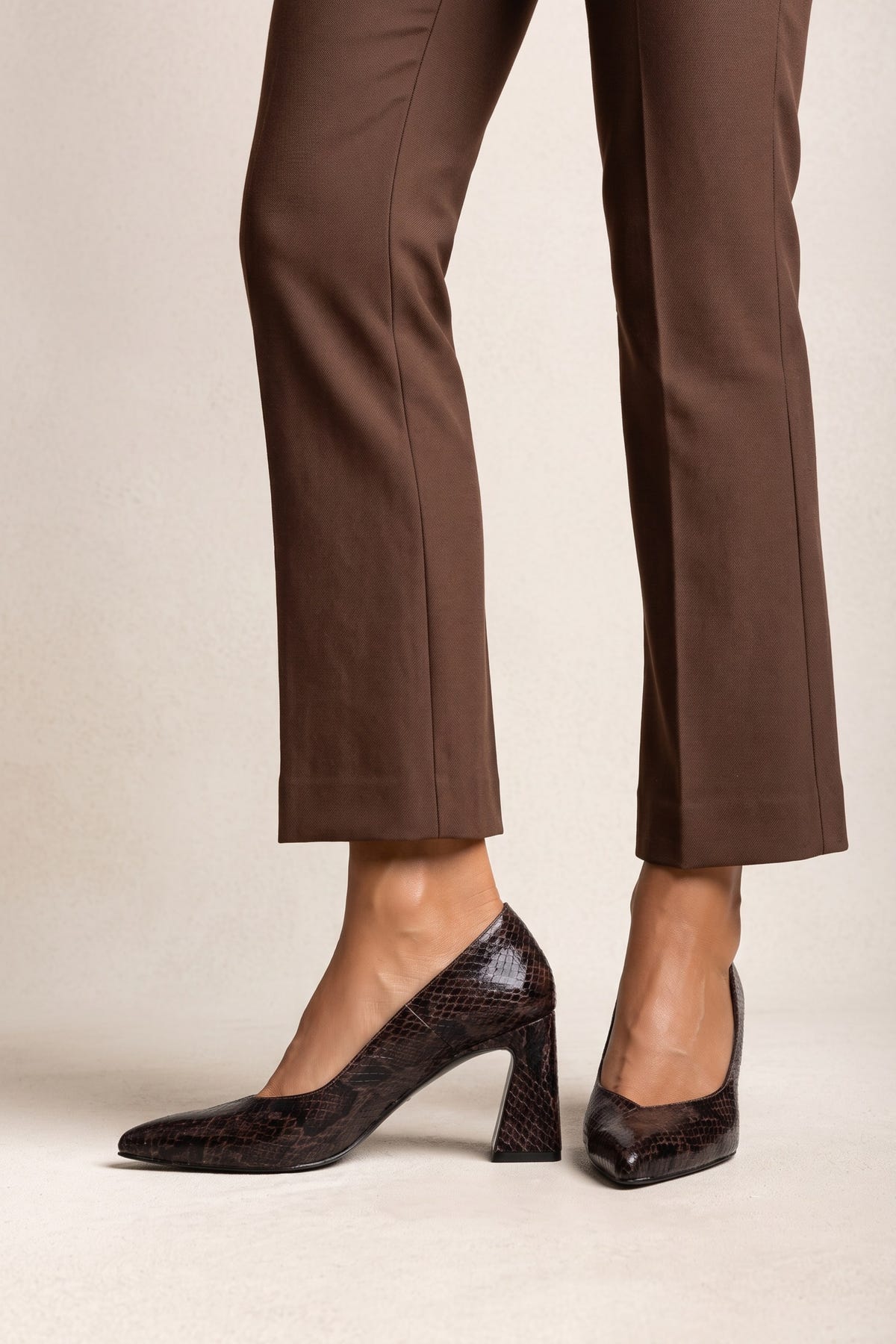 Model in brown trousers wearing brown snake-print pointed-toe pumps standing full length