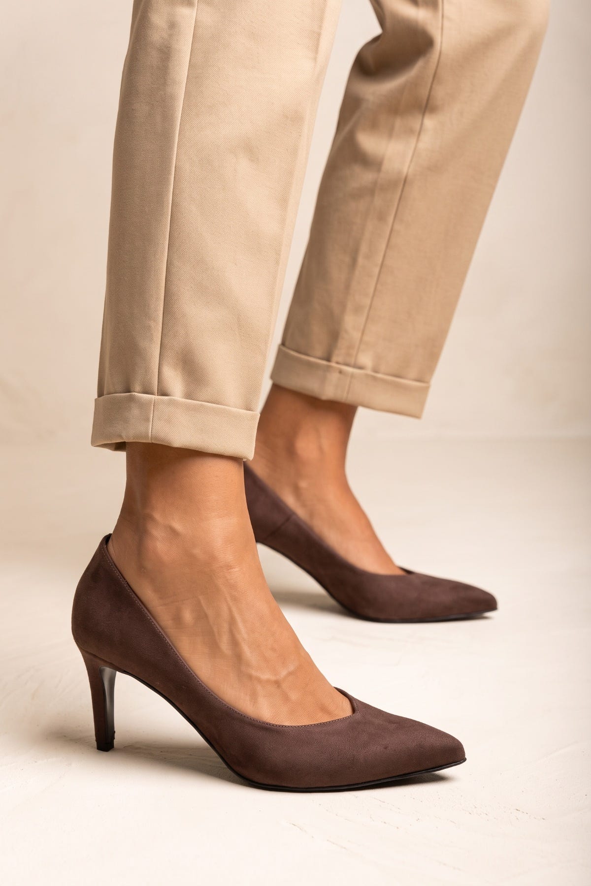 Brown suede pointed toe stiletto pumps with beige cuffed trousers with both feet seen from the side