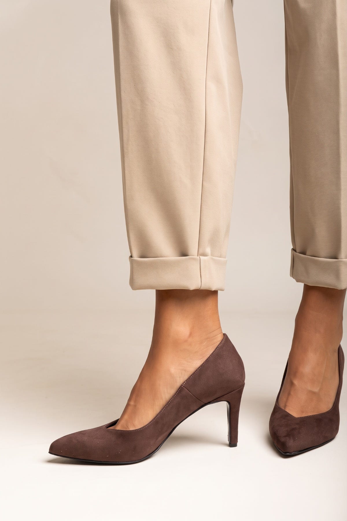 Brown suede pointed toe pumps on model feet below rolled beige trousers seen from the front