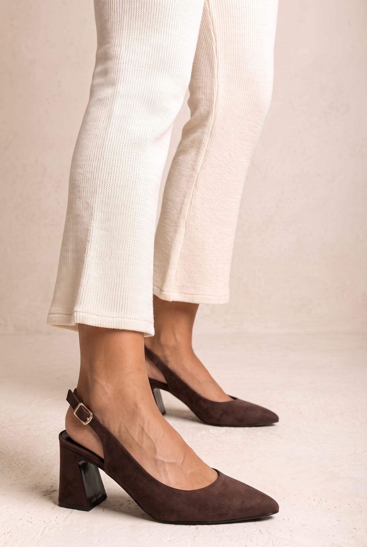 Model wearing brown suede slingback pumps with cream trousers, feet shown from the side against a beige wall