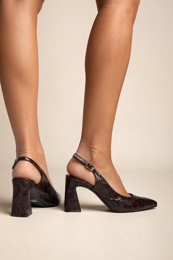 Model wearing brown snake-print slingback pumps with bare legs, seen from behind with the buckled straps visible