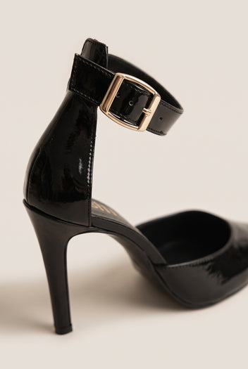 Close-up of a black patent ankle strap with a gold buckle and slim stiletto heel rising from the pointed shoe