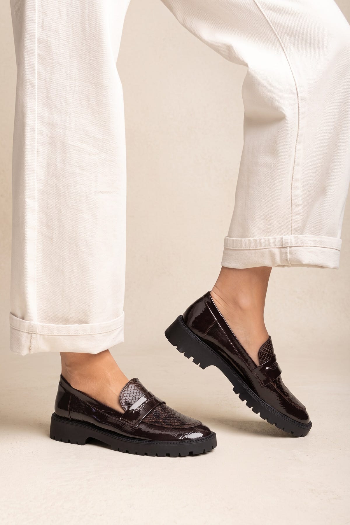 Model wearing brown patent chunky loafers with cream trousers and penny strap, feet angled outward