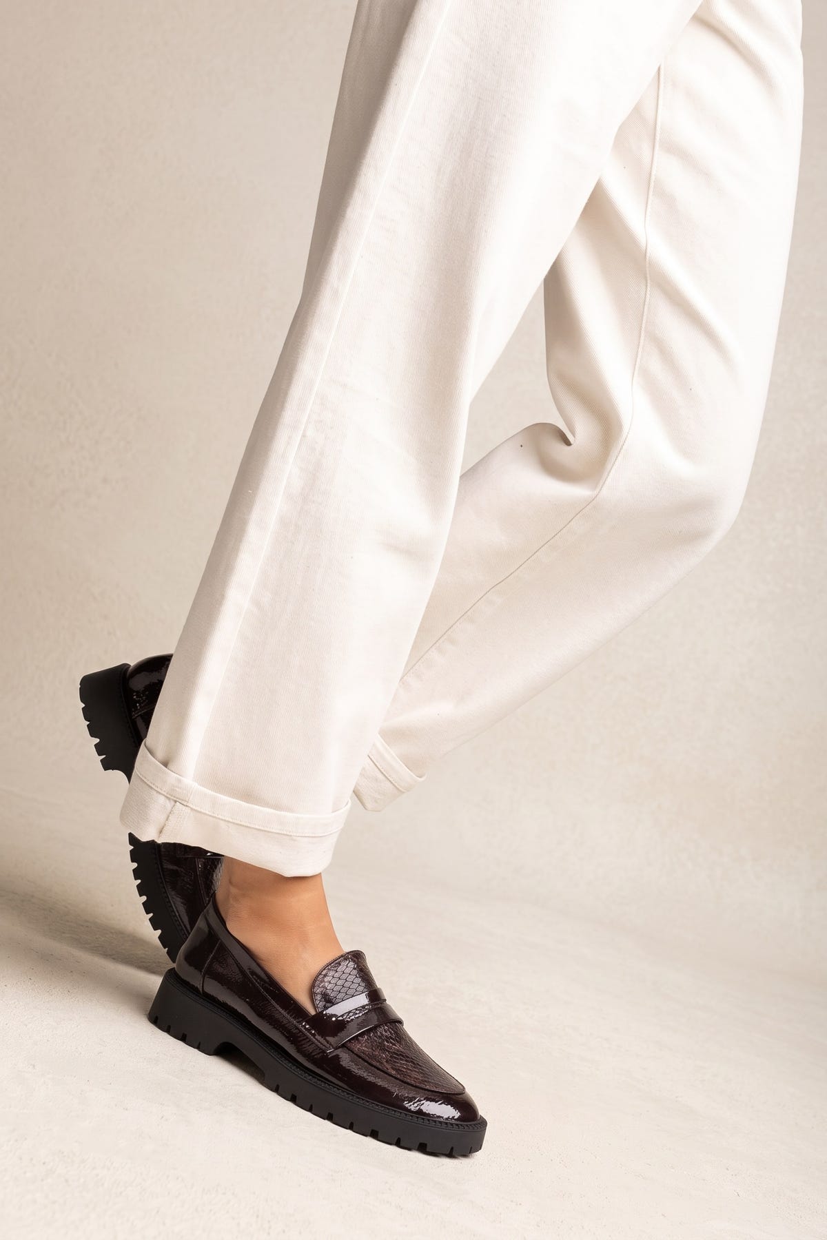 Model wearing brown patent chunky loafers with cream trousers, one foot stepping forward