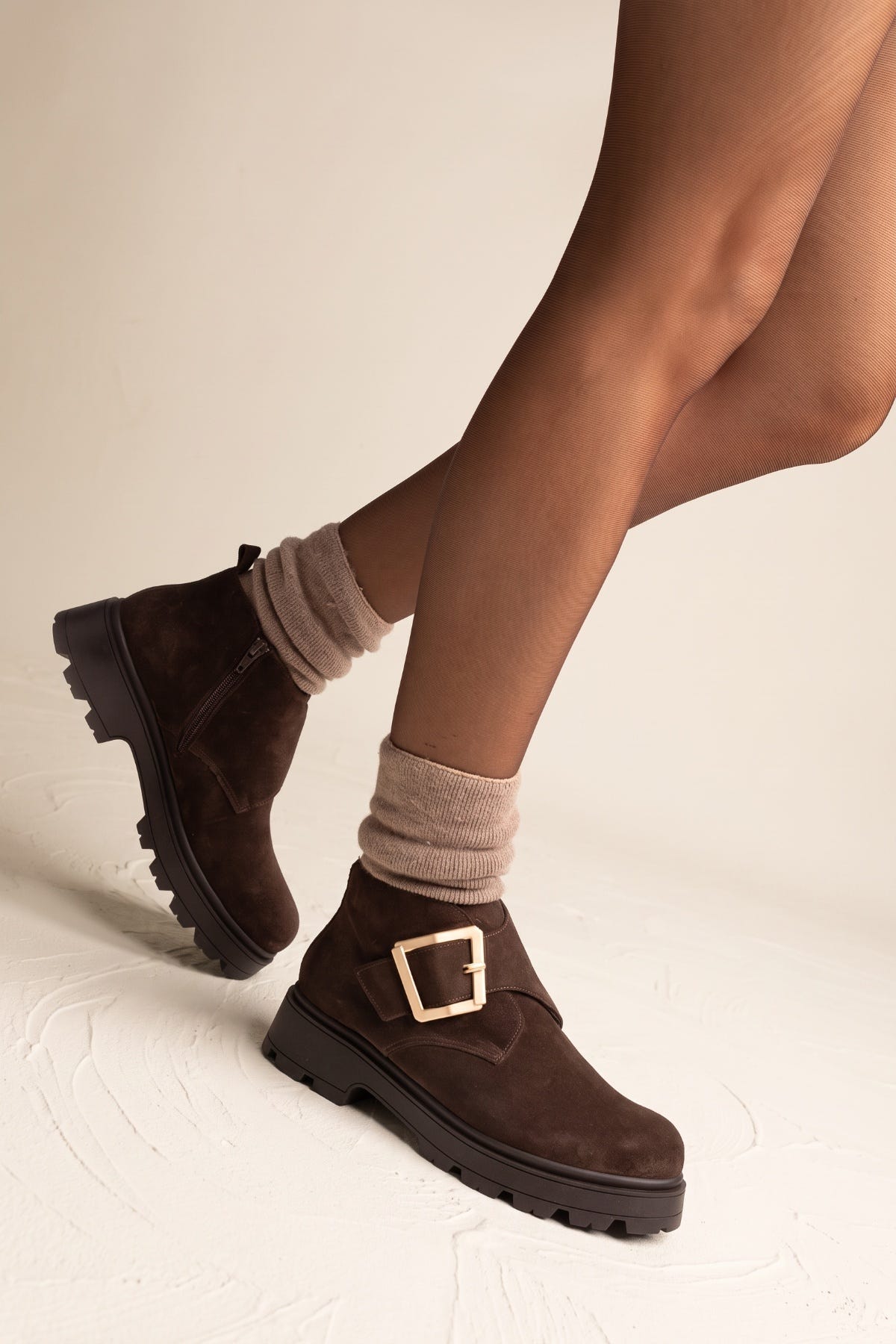 Model with one leg bent wearing brown suede chunky ankle boots with gold buckle and knit socks