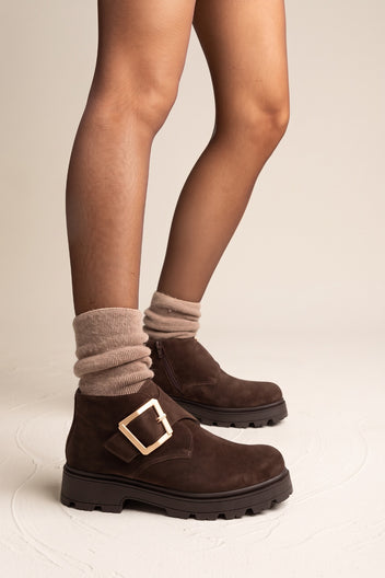 Model standing with feet apart in brown suede ankle boots with gold buckle and knit socks