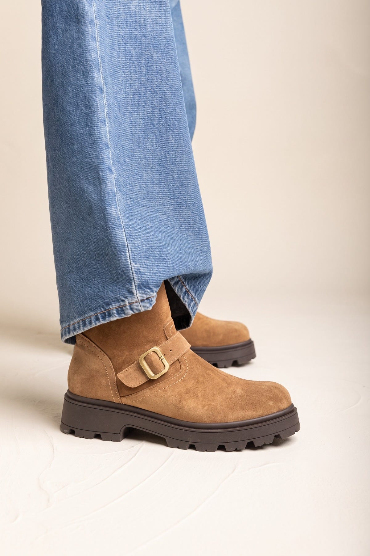 Taupe suede biker ankle boots with ankle buckle strap and dark lug sole, front view of both feet in wide-leg jeans