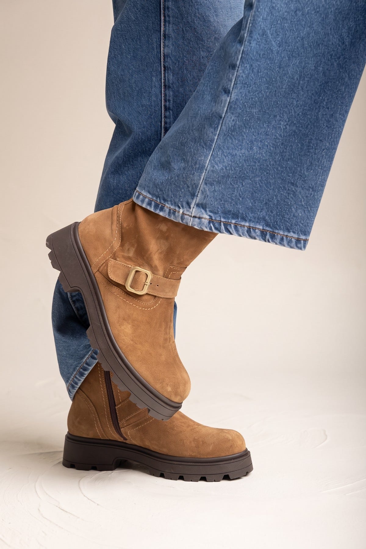 Taupe suede biker ankle boot with buckle strap and dark brown lug sole, close side view of foot in rolled jeans