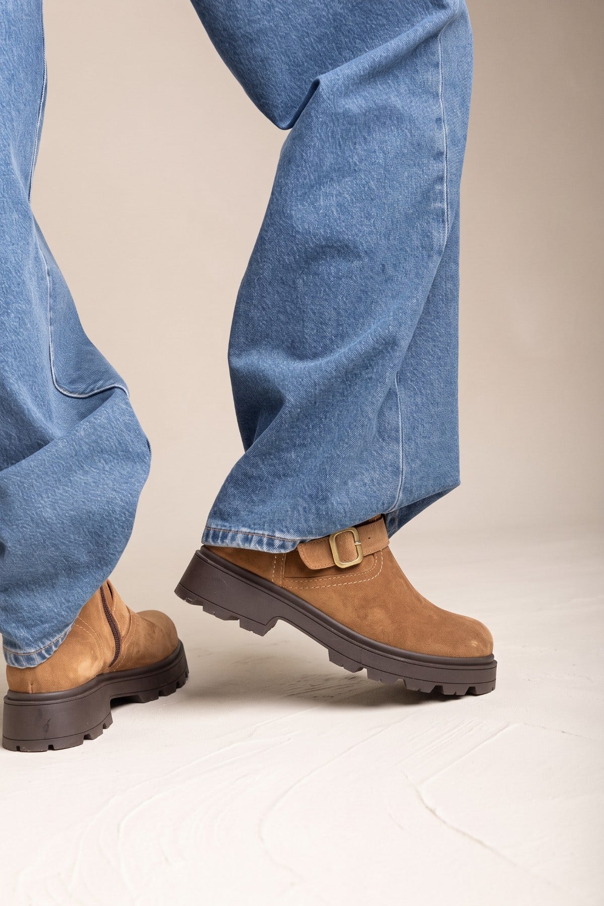 Model wearing taupe suede biker ankle boots with buckle strap and thick dark lug sole, side view with blue jeans
