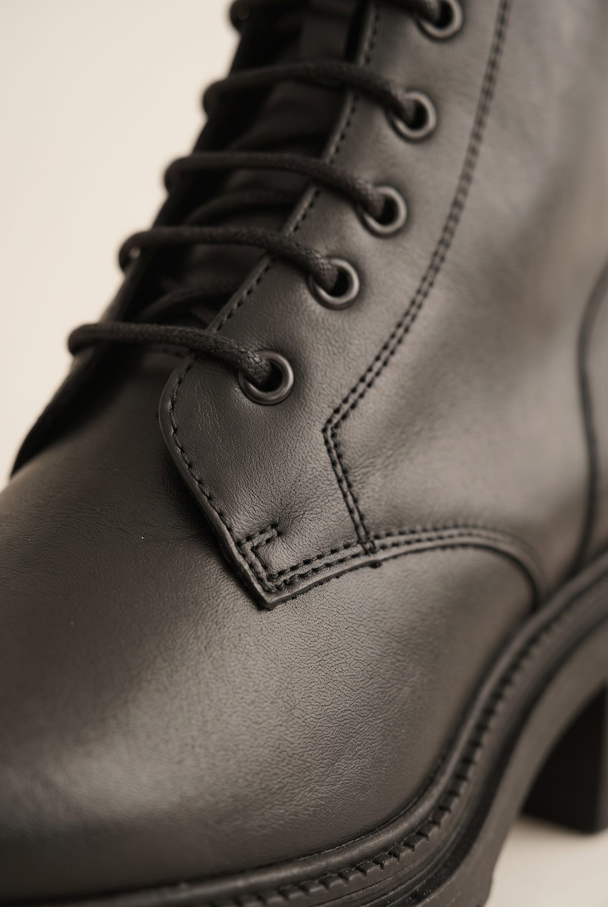 Close-up of a black leather lace-up boot with black eyelets, laces and contrast stitching above a chunky lug sole