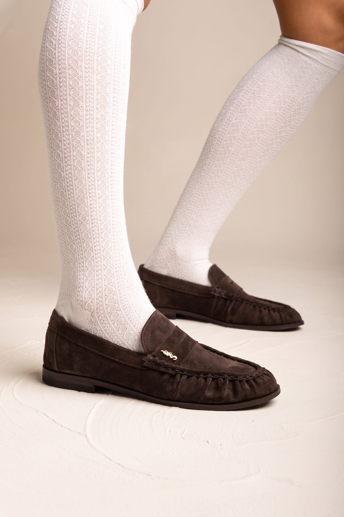 Brown suede flat loafers with pleated detail and small gold accent worn with white knee socks