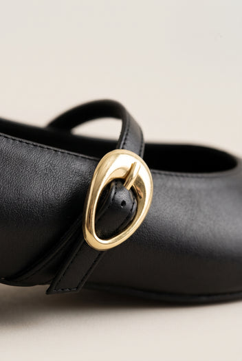 Close-up of black leather Mary Jane shoe with strap and oval gold buckle, textured smooth leather