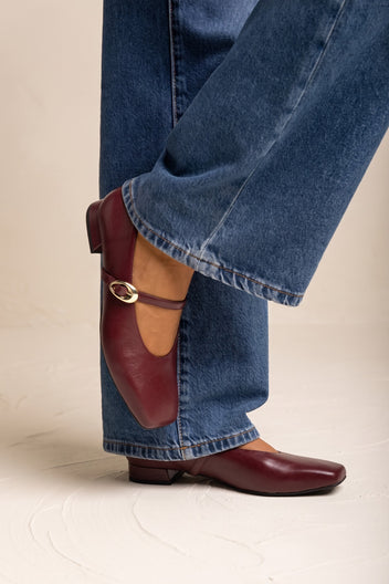 Bordeaux matte square toe flat with ankle strap and metal buckle, side view on model in jeans