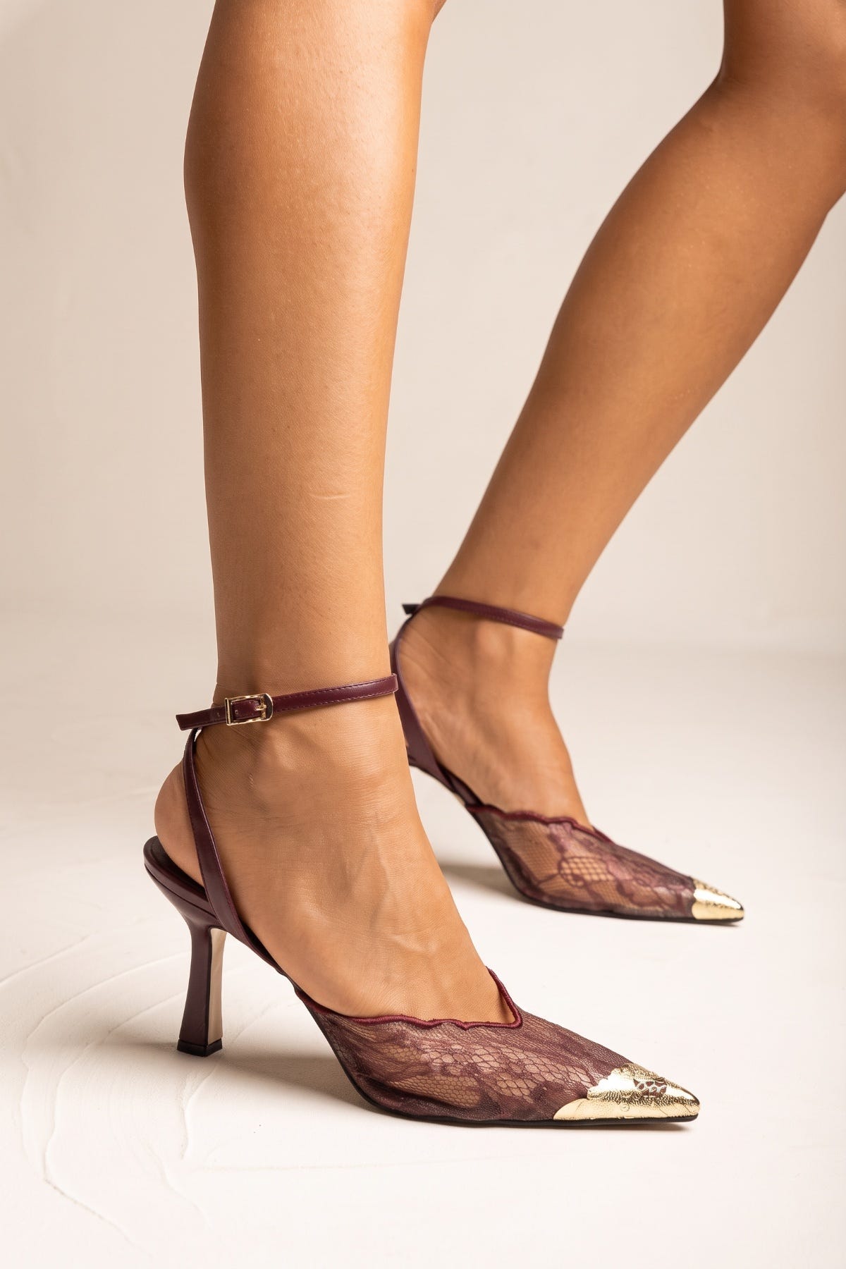 Model wearing bordeaux lace stiletto pumps with gold pointed toe caps and thin ankle straps