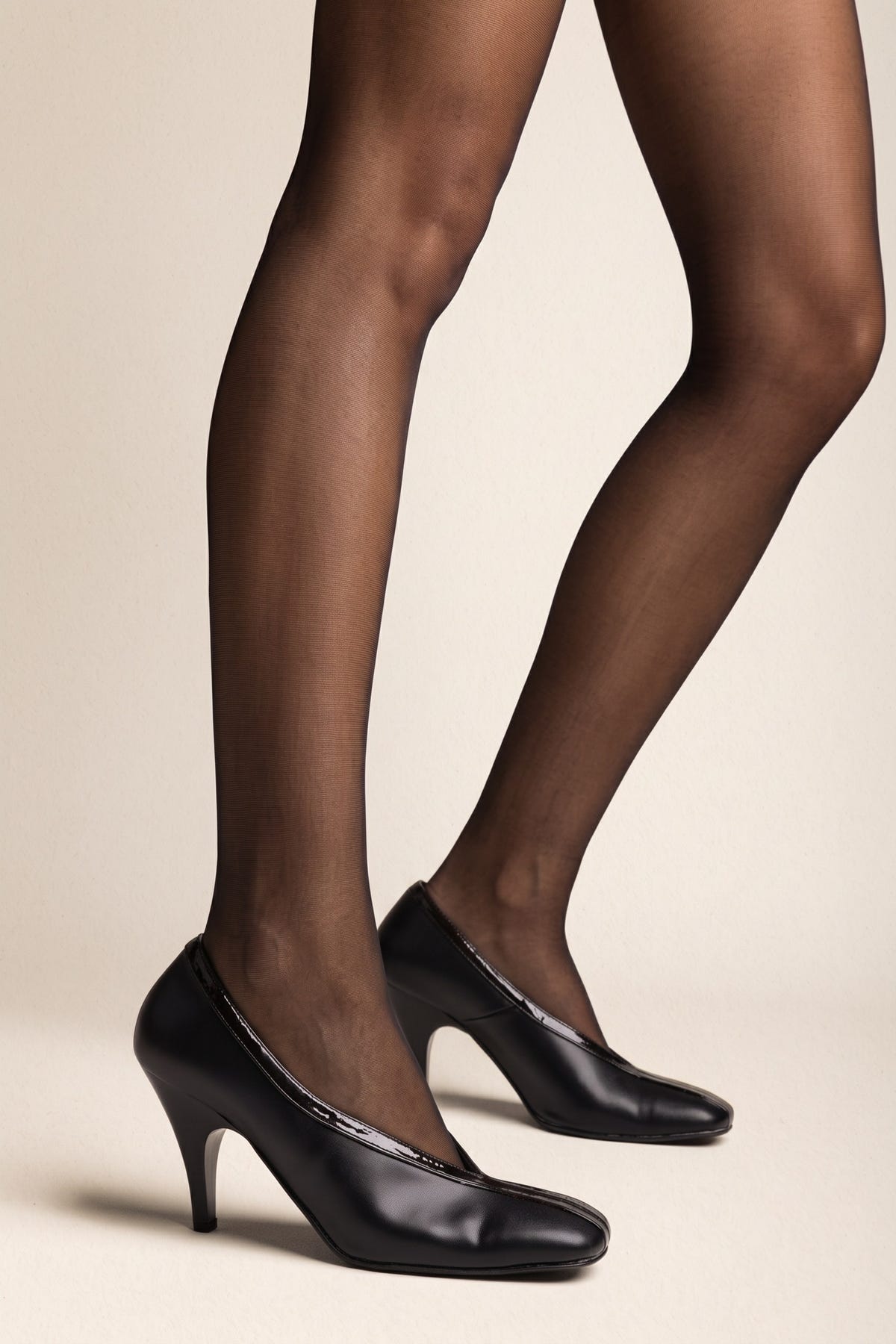 Black smooth leather pump with rounded square toe, patent trim along the topline and a high slim heel, side view worn on model