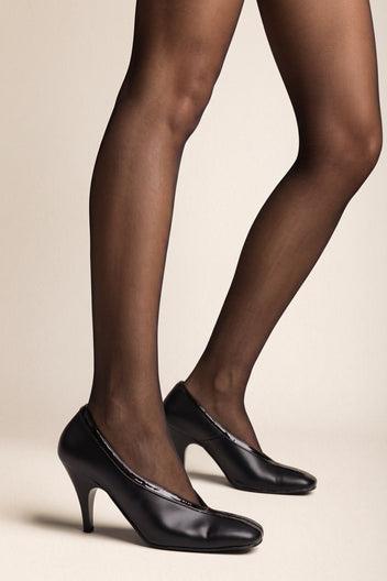 Black smooth leather pump with rounded square toe, patent trim along the topline and a high slim heel, side view worn on model