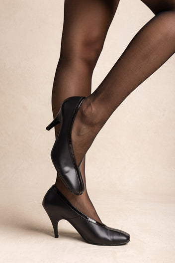 Black smooth leather pumps with a rounded toe, thin stiletto heel and glossy trim along the opening, on model