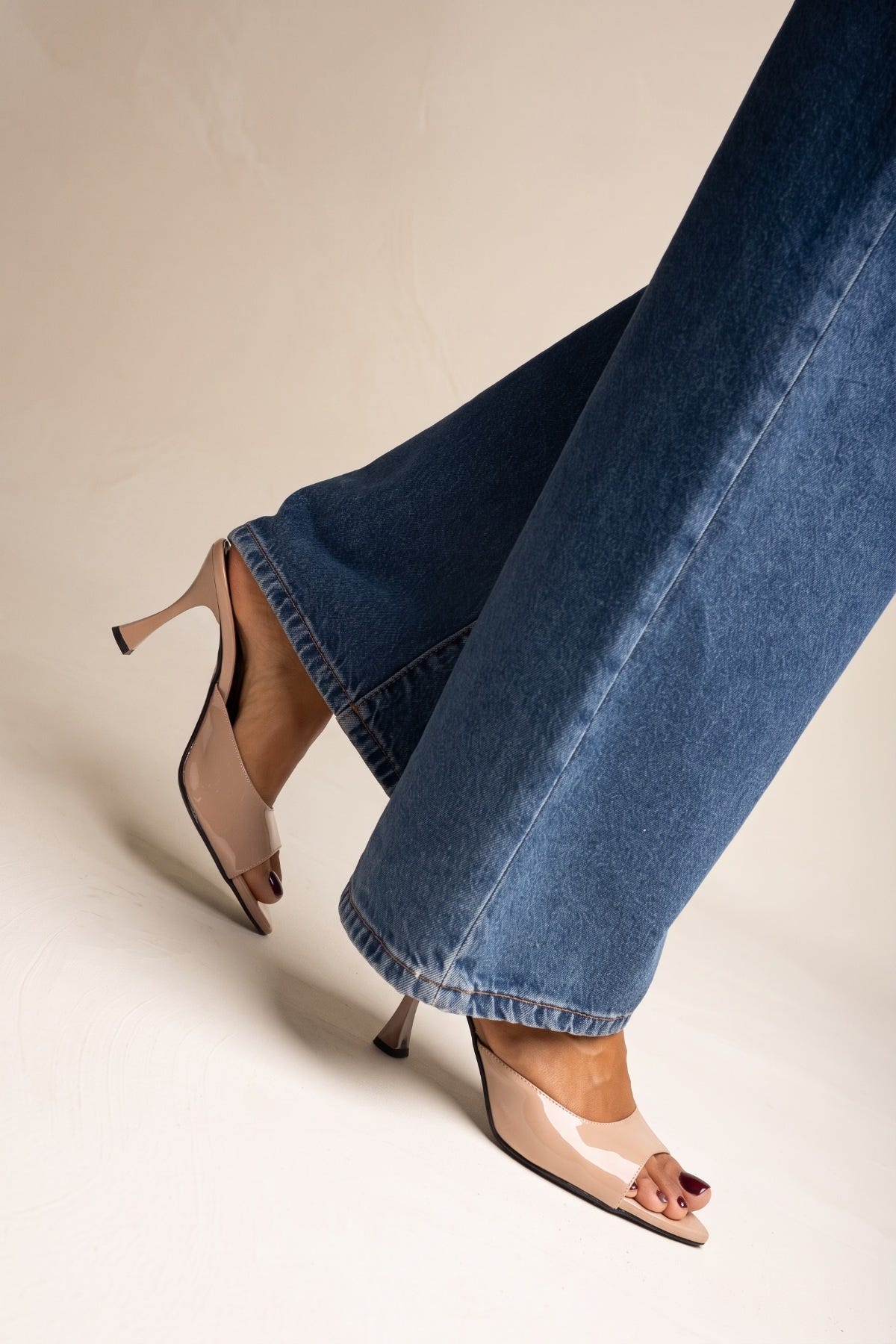 Nude patent stiletto mule with clear open toe and thin heel, side close view on model in jeans