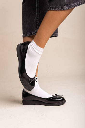 Model wearing black patent ballet flats with bow detail and round toe, raised leg view with white ankle socks and dark cropped trousers