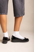 Model wearing black patent ballet flats with a small bow on the vamp and thin sole, side view with cropped trousers and white socks