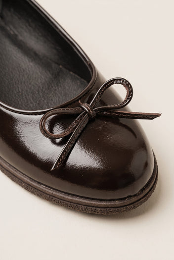 close-up of a glossy dark brown patent ballet flat toe with a thin bow detail and rounded cap toe