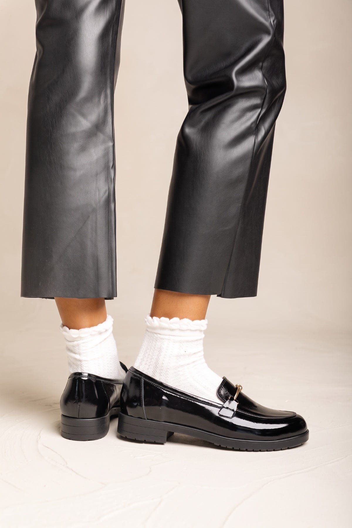 Lower legs of a model in black trousers wearing black patent horsebit loafers with white ruffled socks