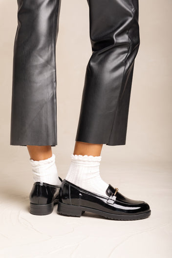 Lower legs of a model in black trousers wearing black patent horsebit loafers with white ruffled socks
