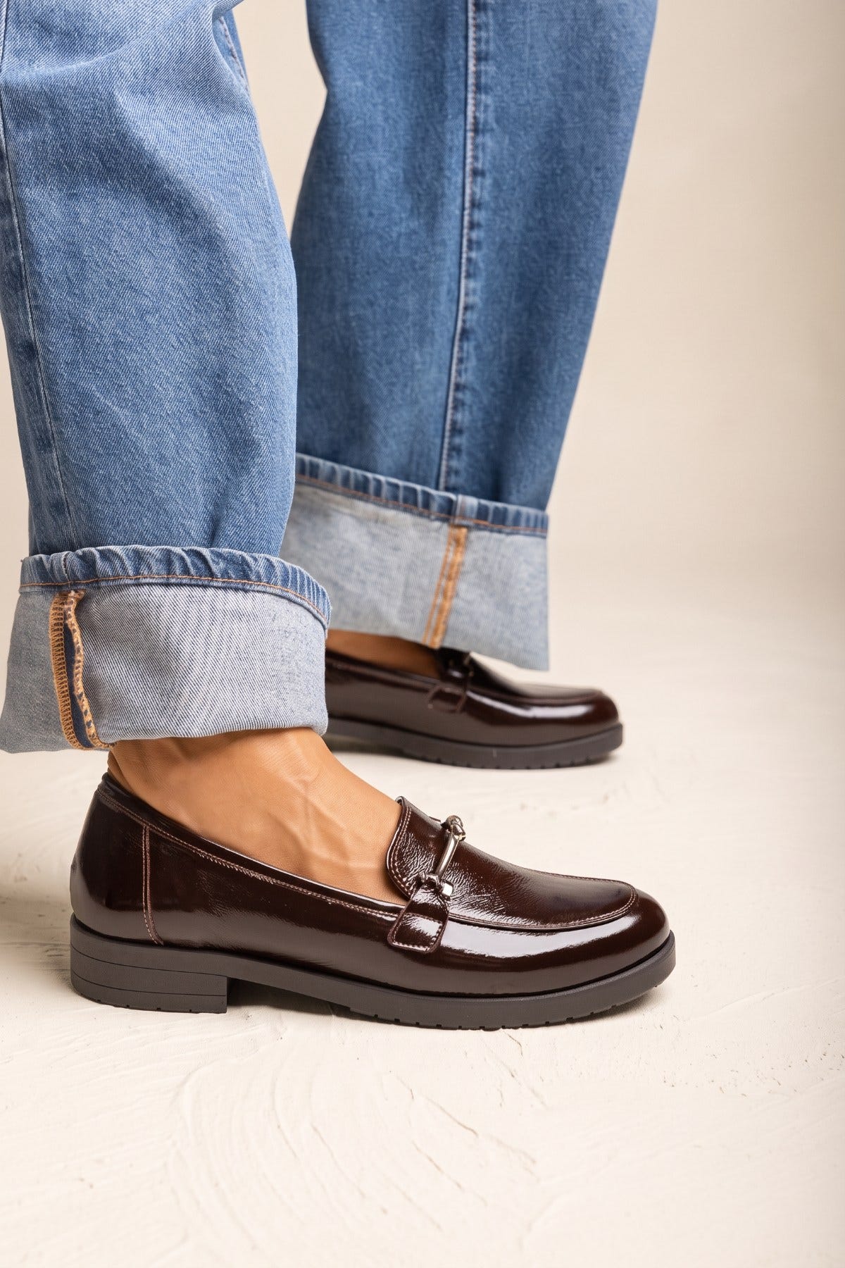 Model in cuffed blue jeans wearing brown patent horsebit loafers with a chunky dark sole standing view