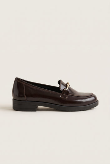 Side view of a single brown patent horsebit loafer with a gold metal bit and low block heel on a dark sole