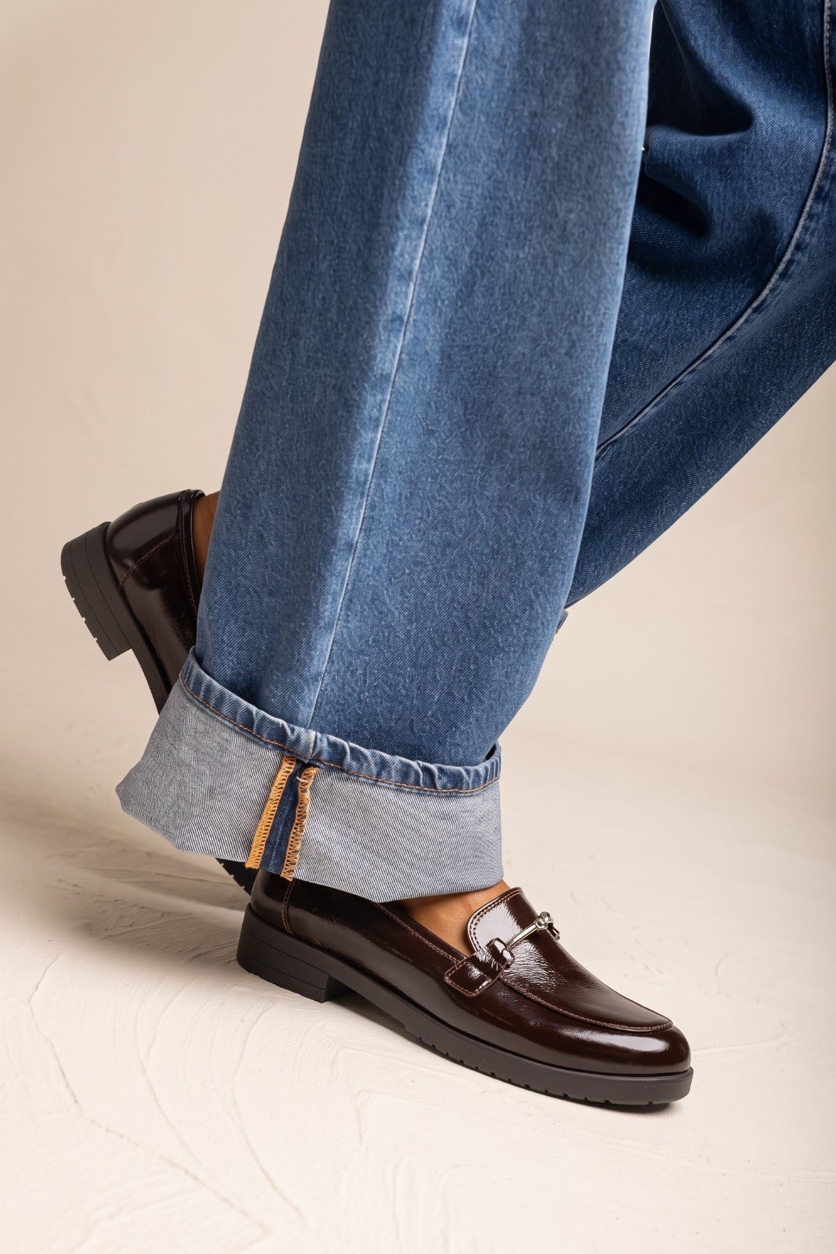Cropped view of model in cuffed jeans with a brown patent loafer on foot and dark sole at the heel