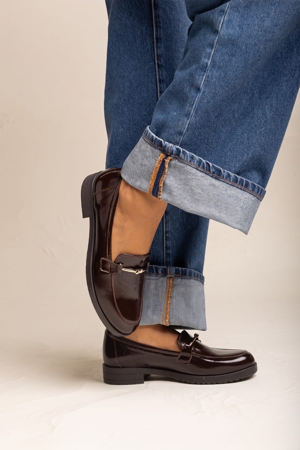 Model in cuffed jeans with brown patent horsebit loafers side angle showing the gold bit at the vamp