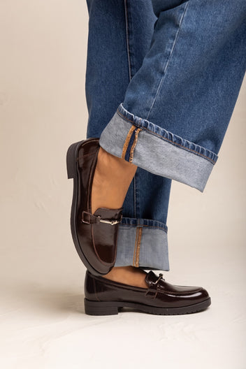 Model in cuffed jeans with brown patent horsebit loafers side angle showing the gold bit at the vamp
