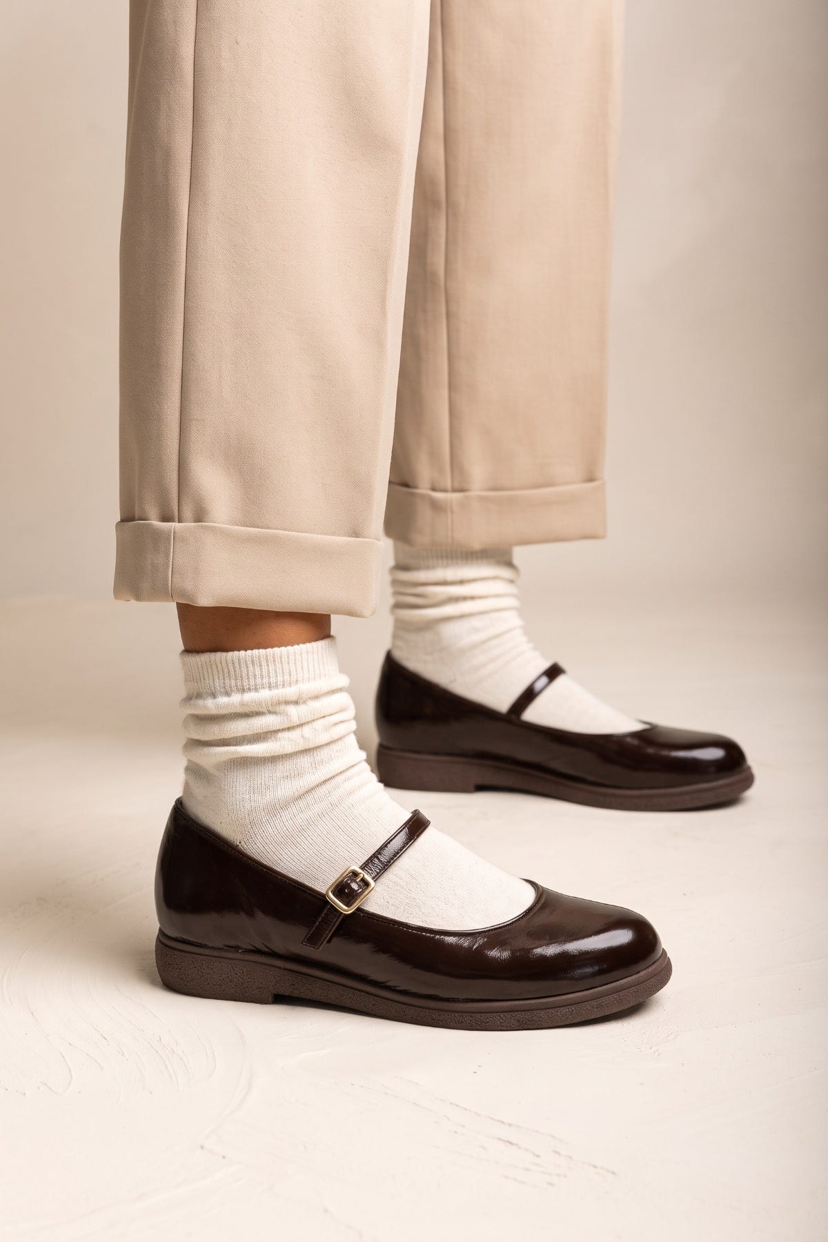 Dark brown patent Mary Jane flats with ankle strap, worn with white socks and beige cuffed trousers