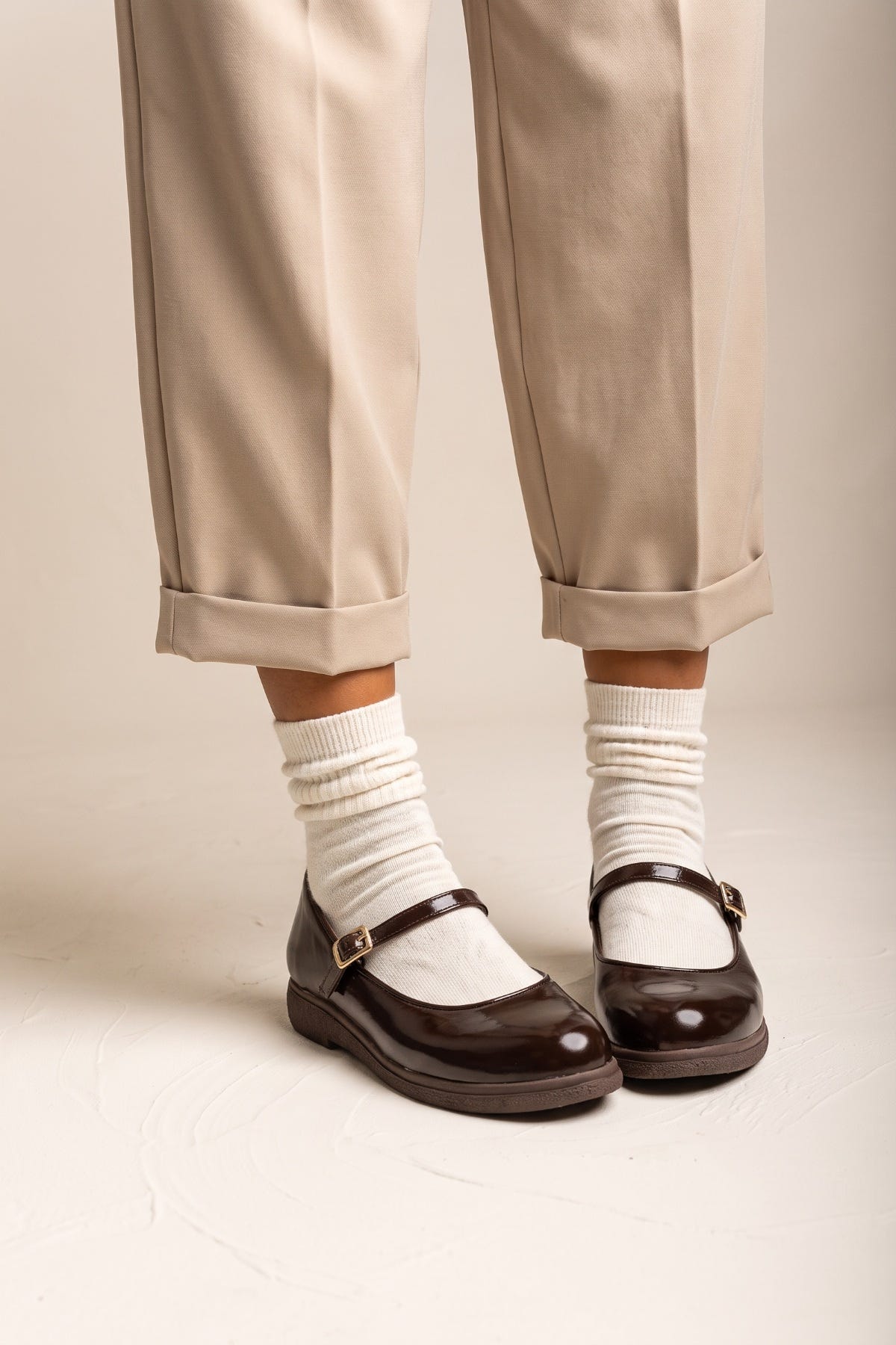 Pair of dark brown patent Mary Jane flats with ankle straps, white socks and beige trousers, side view