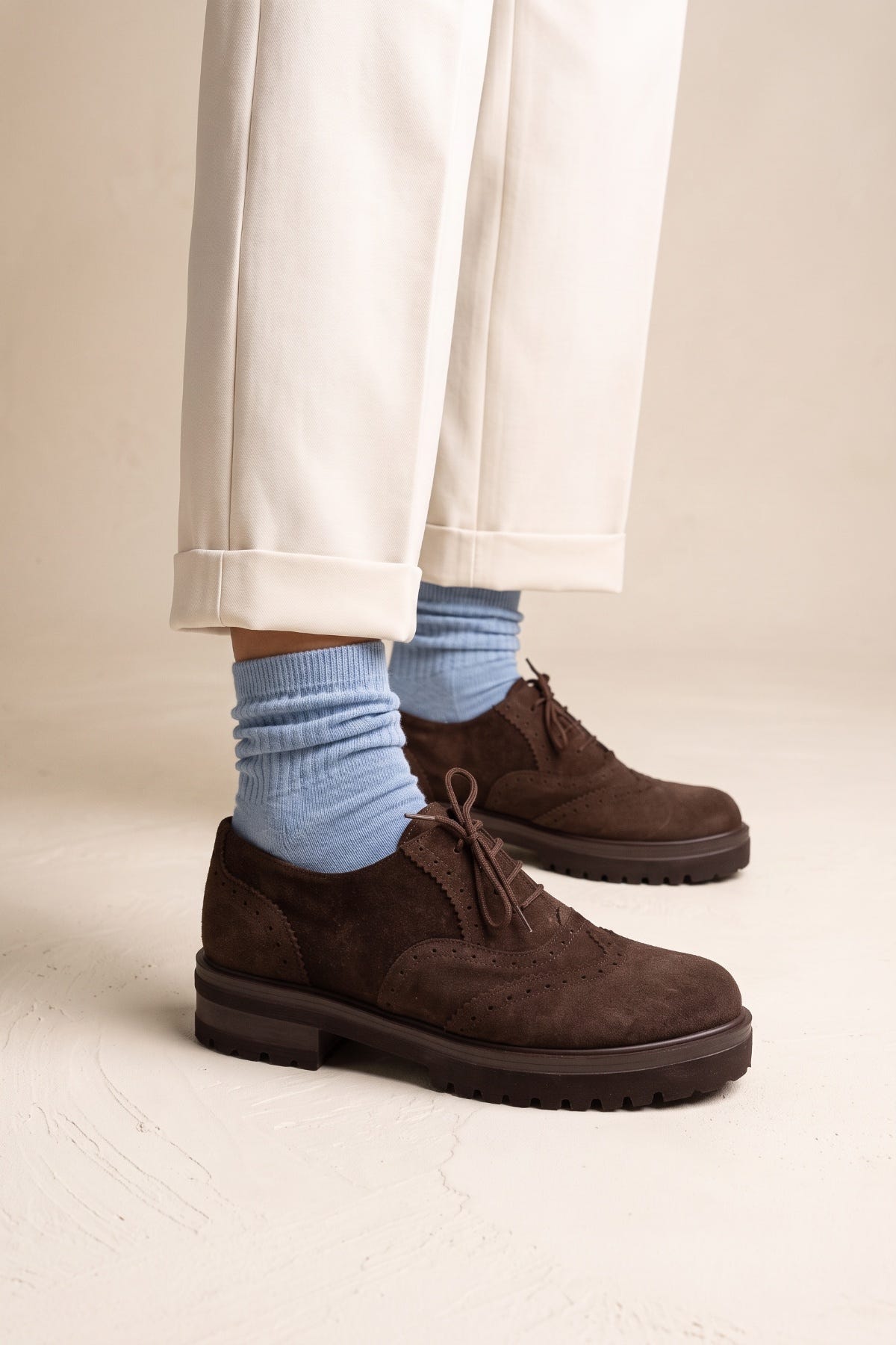 Side view of model wearing brown suede brogue lace ups with chunky rubber sole and light blue socks beneath cream cuffed trousers